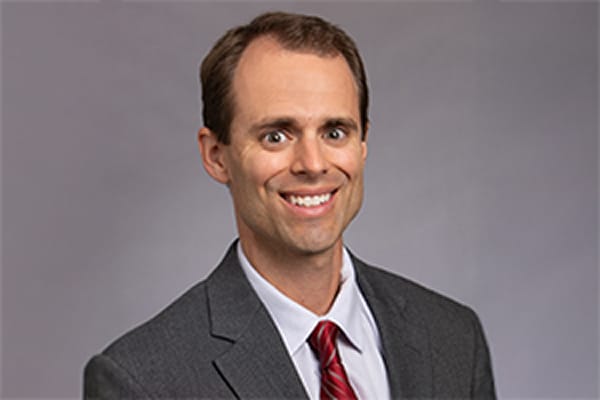 Smiling man in a suit and red tie against a gray background.