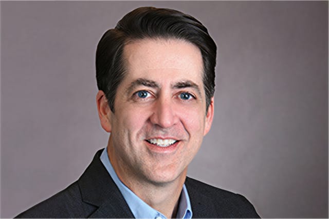 Smiling man in a professional suit with a gray background.
