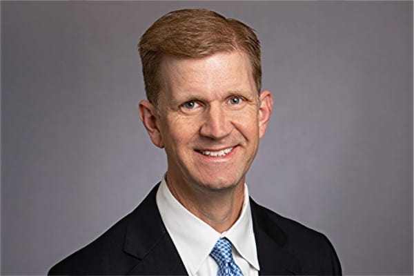 Smiling man in a suit with a blue tie against a gray background.